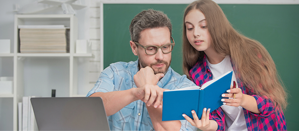 Un professore seduto davanti al computer indica qualcosa sulla pagina di un libro tenuto da una sua alunna adolescente